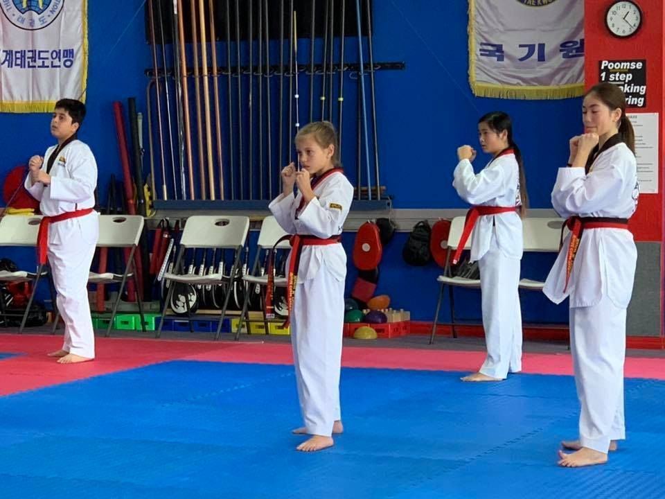 Four people in white taekwondo uniforms and red belts stand in a line on a blue mat inside a studio.