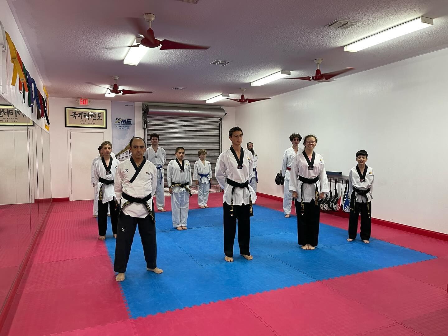 A group of students in white martial arts uniforms with black belts stand in rows on a blue mat in a gym.