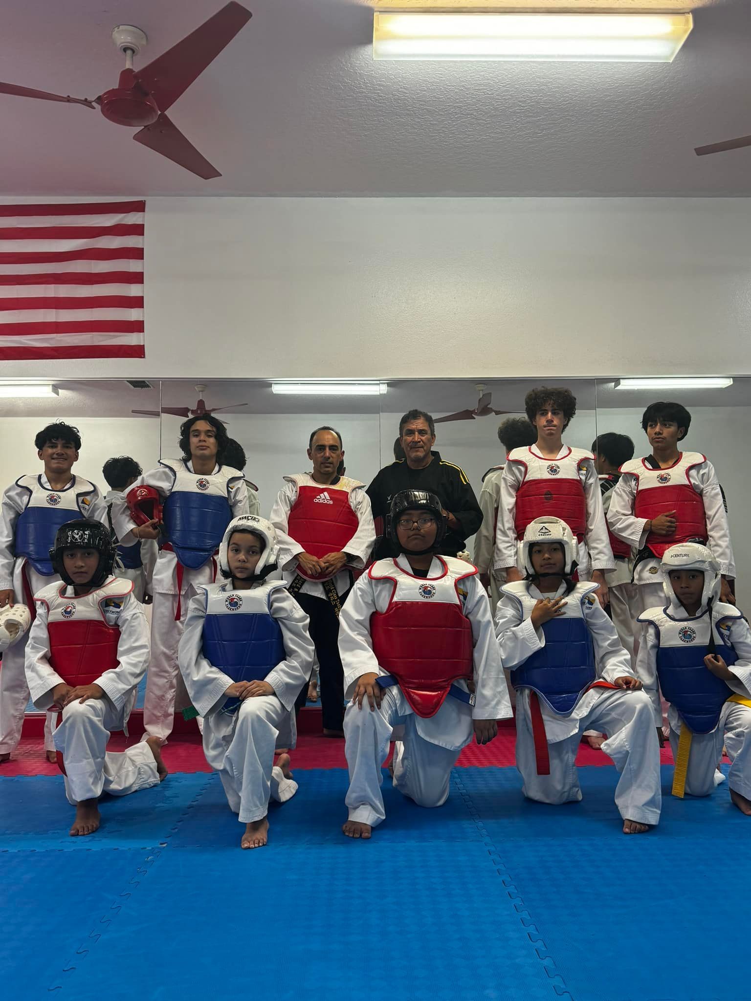 A group of martial artists in white uniforms and chest protectors kneeling and standing in a studio with a US flag.