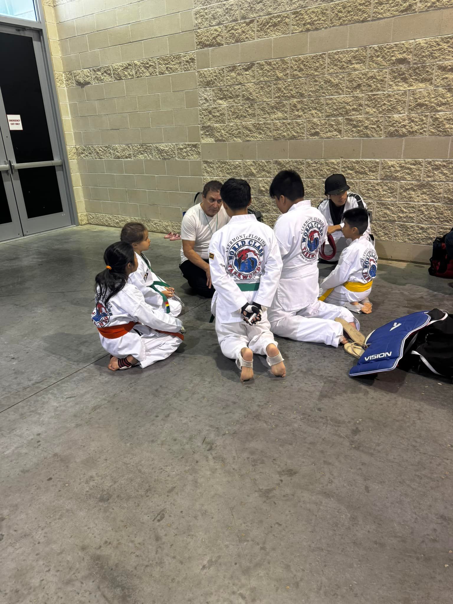 Students in taekwondo uniforms kneel on a gym floor, listening to an instructor near a brick wall.
