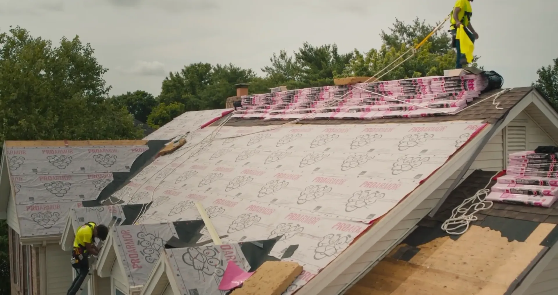 Roofers on a house roof installing shingles. Pink underlayment and equipment visible. Overcast day.