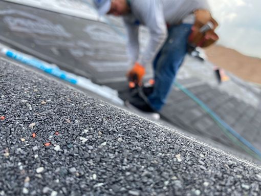 Roofer installing shingles on a roof; blue jeans, orange gloves, and a tool belt are visible.
