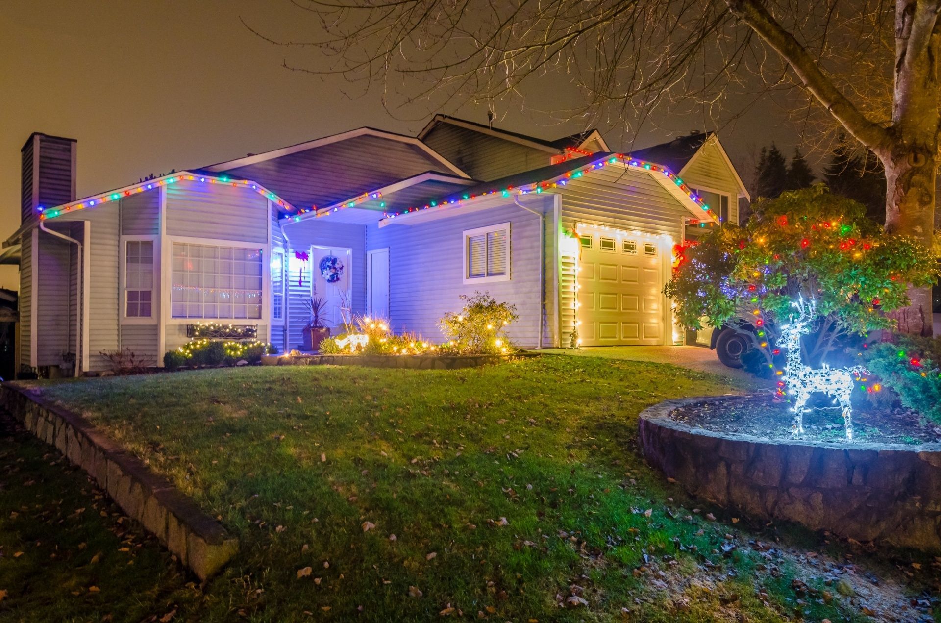 House decorated with Christmas lights at night, glowing along roofline, shrubs and garage.