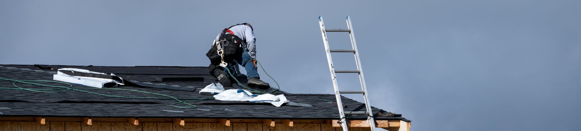 Roofer on a dark roof near a ladder under an overcast sky.
