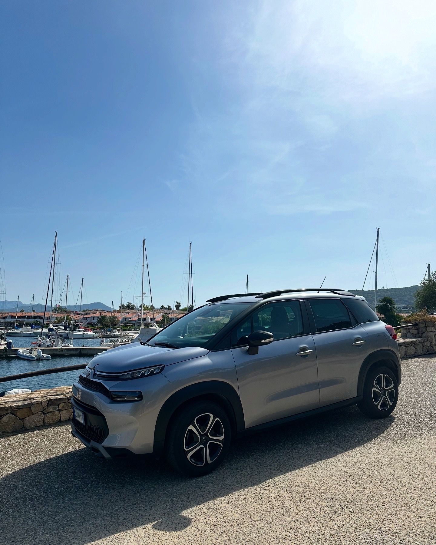 Citroen C3 Aircross argentata parcheggiata vicino a un muro di pietra, con porto e cielo azzurro sullo sfondo.