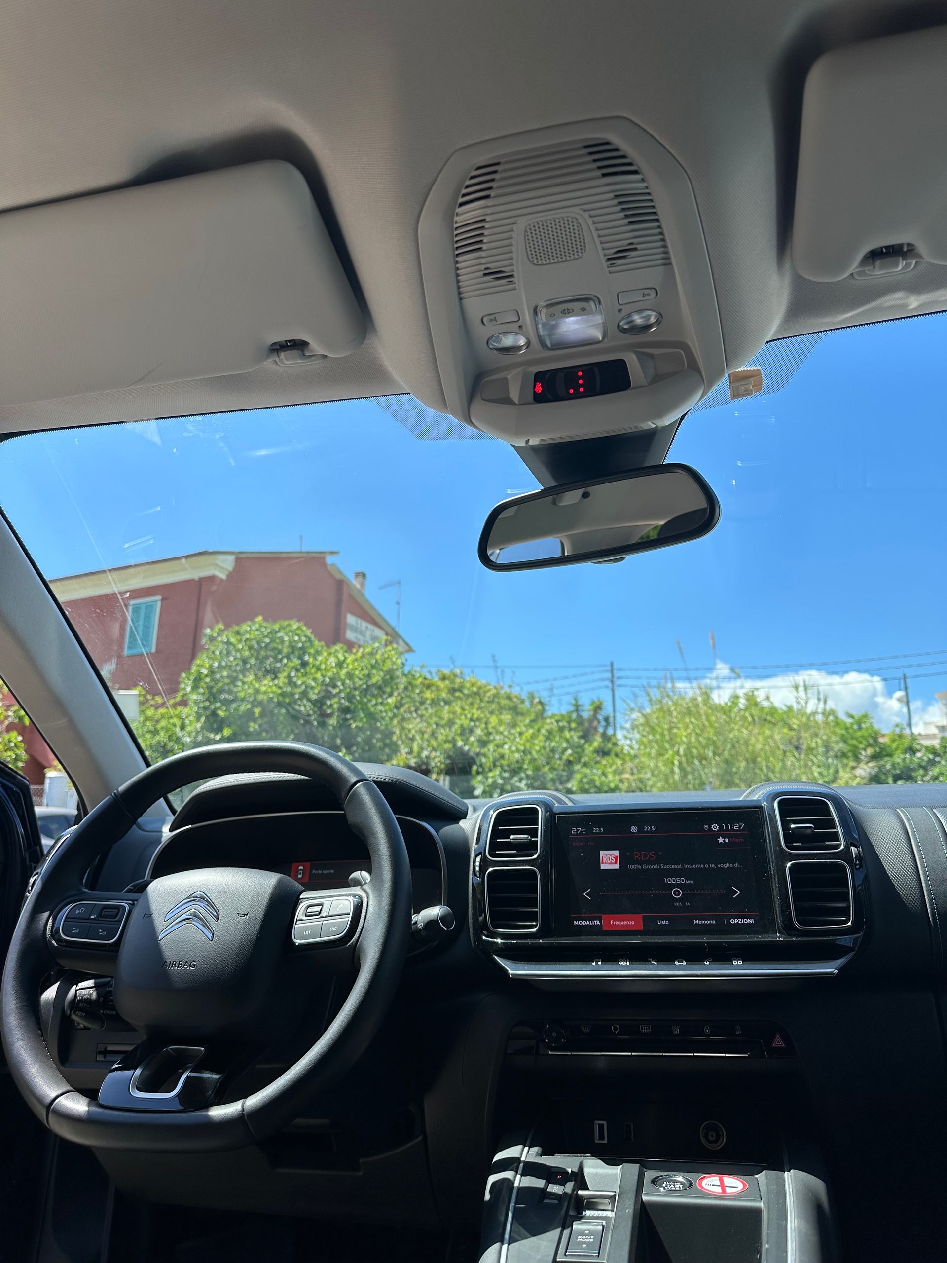 Vista interna di un'auto: cruscotto, volante, console superiore e parabrezza con vista esterna di un edificio e cielo azzurro.