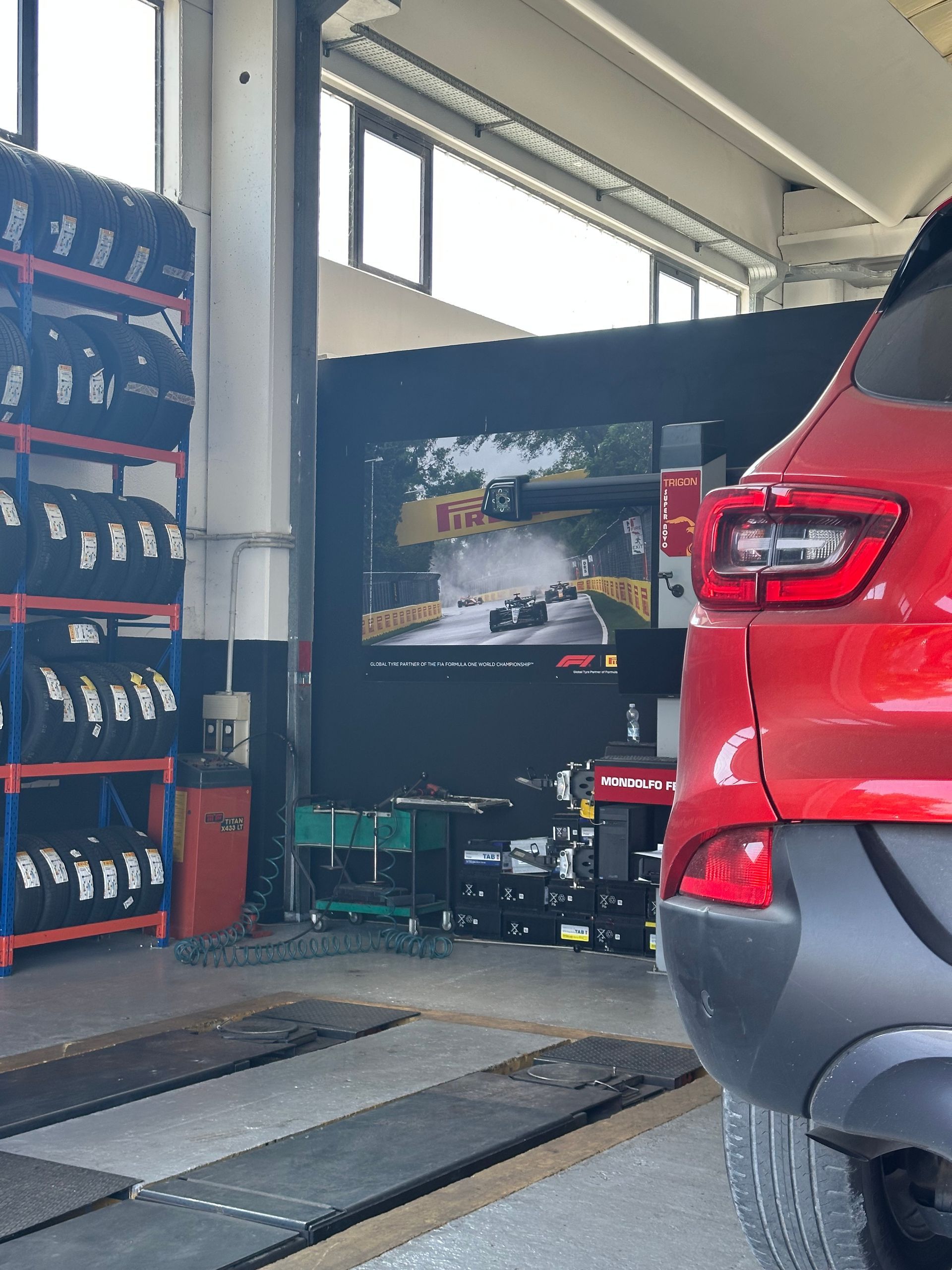 Un'auto rossa in un'autofficina, pneumatici accatastati sugli scaffali, uno schermo che mostra la Formula 1.