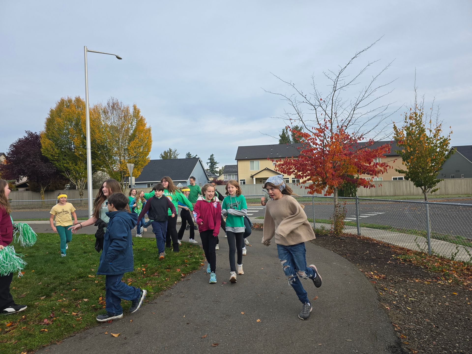 A group of people, many wearing green, running down a paved path outdoors. Fall trees in the background.
