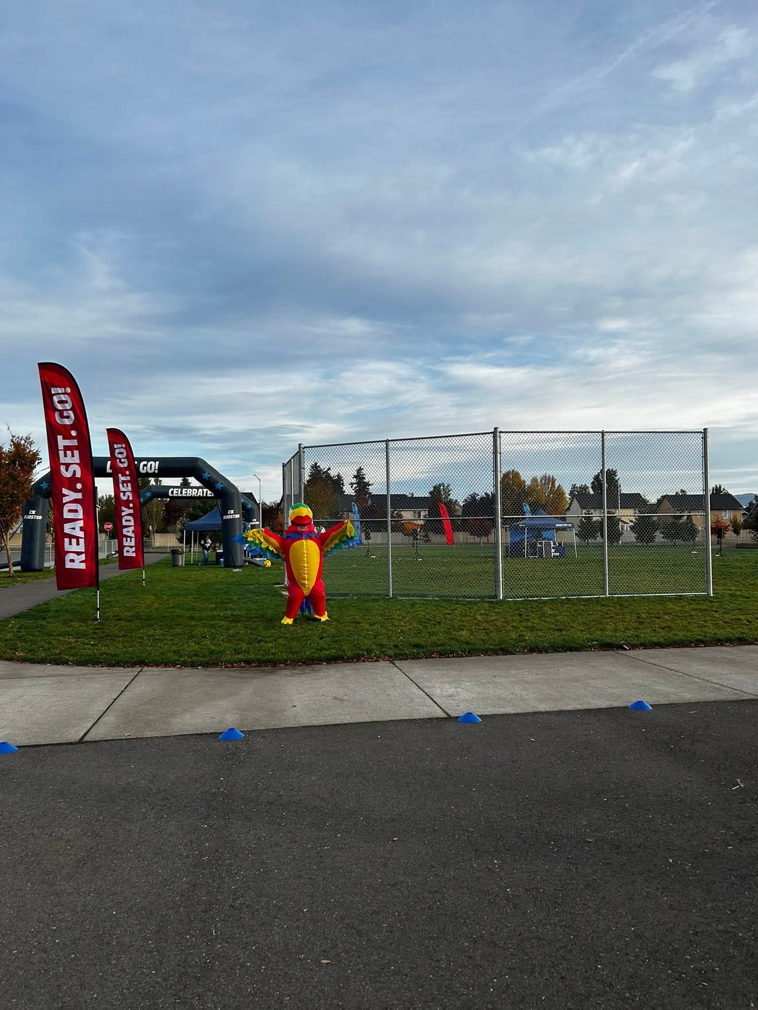 A person in a dragon costume stands on grass near a fenced-in obstacle course; two red banners read