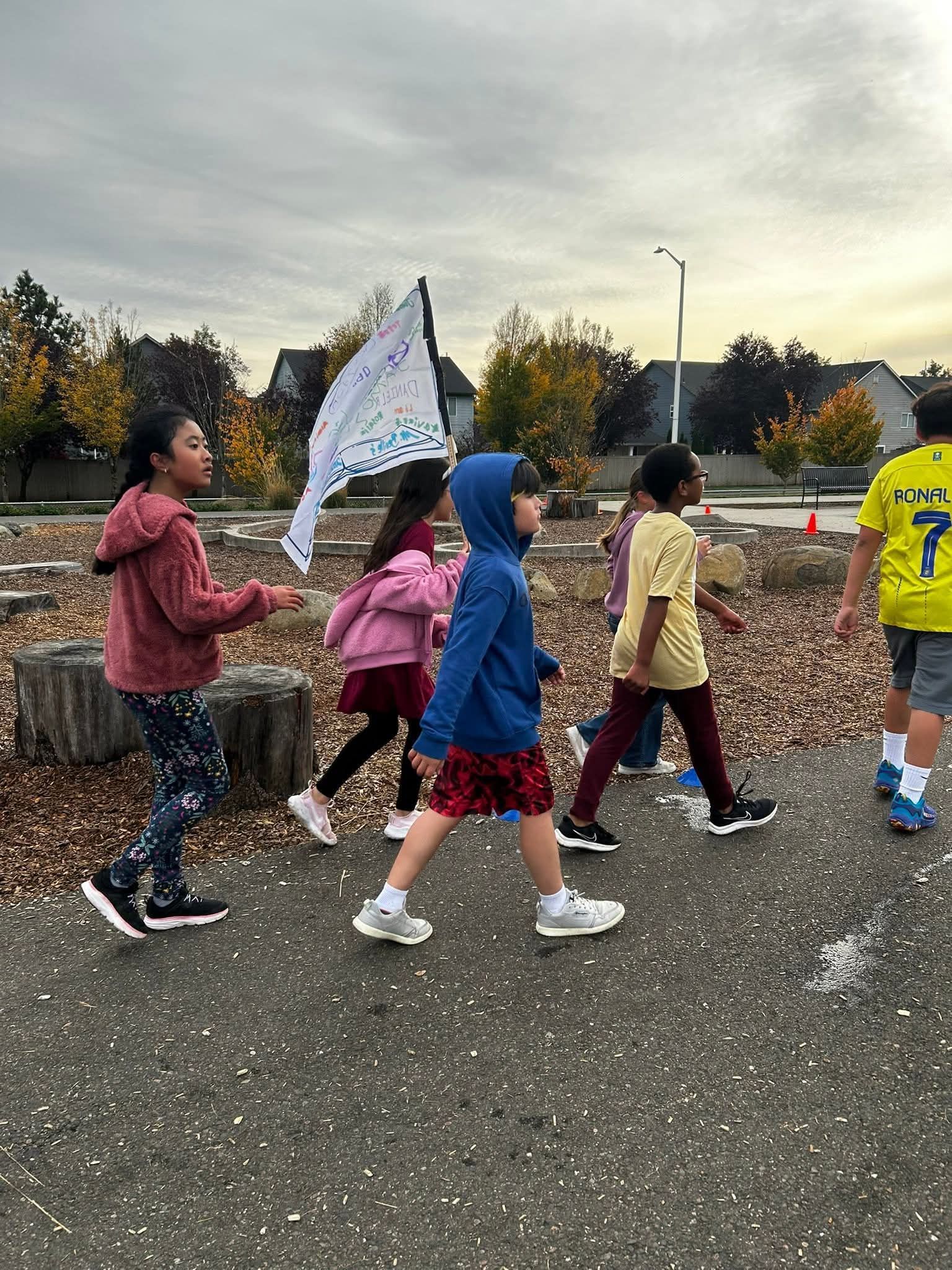 Children walking on a path, one holding a flag. Overcast sky, trees in the background.