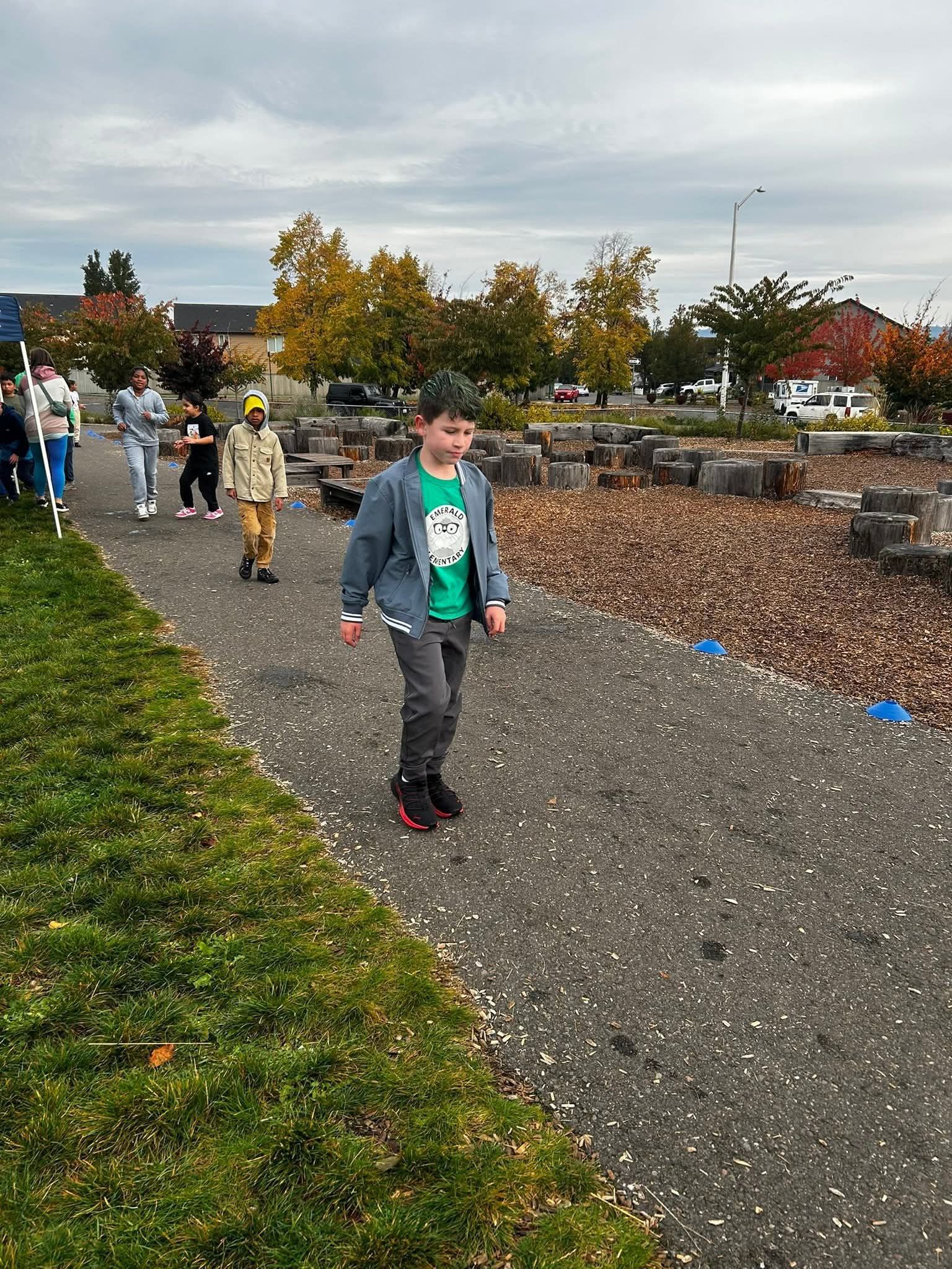 Boy in green shirt and jacket walks on a path; others behind. Fall trees and open area.