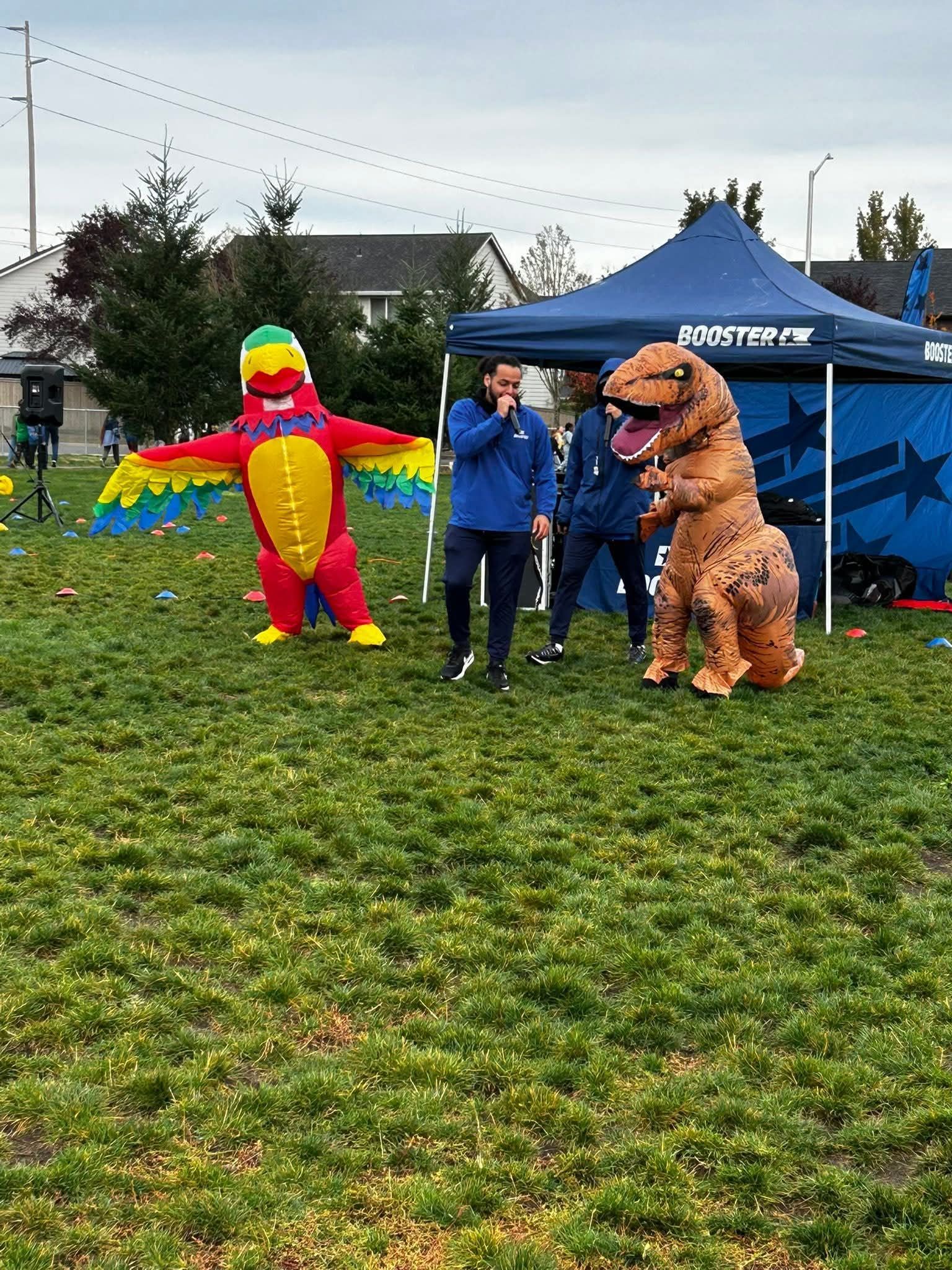 People in blue uniforms talking to a parrot and a dinosaur mascot in a grassy field. A tent is behind them.