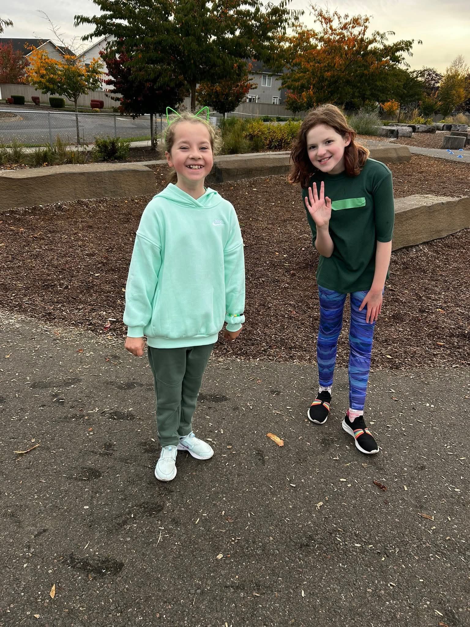 Two girls smiling outdoors, one in green sweatsuit, the other in a green shirt and patterned leggings, waving.