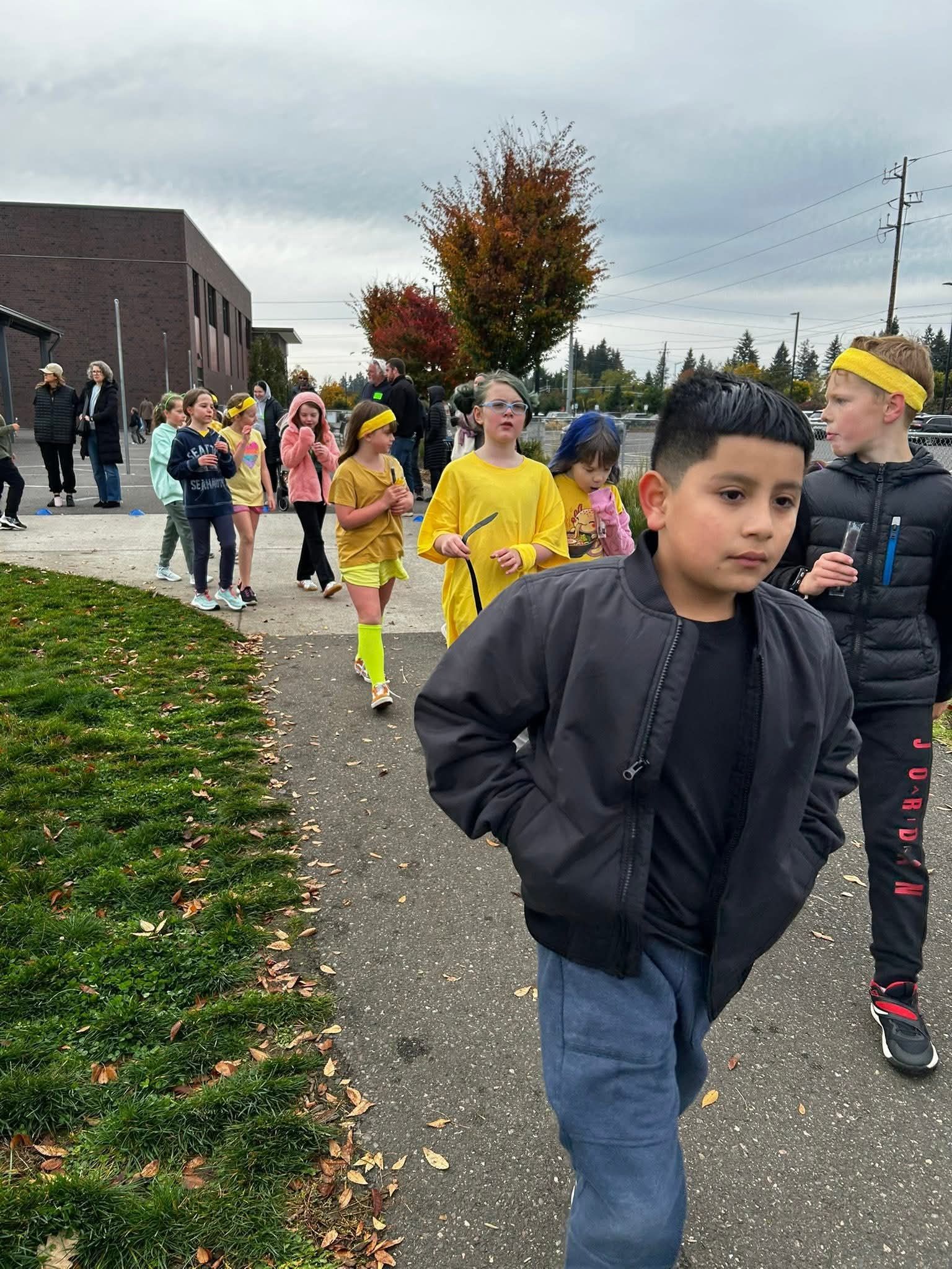 Children walking on a path, some wearing yellow. Cloudy day, school building in background.