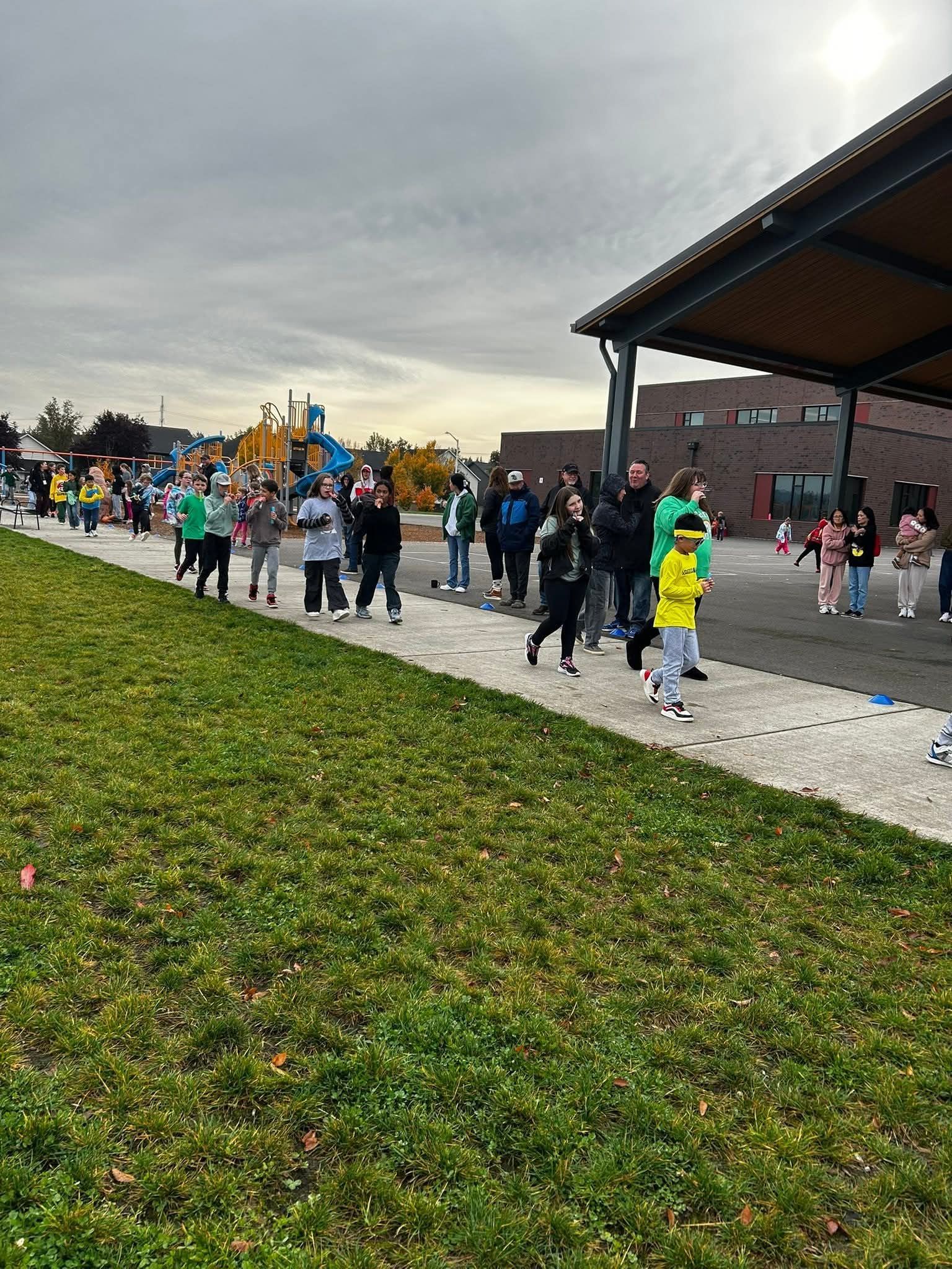 Children walk on a path at school, toward a covered area. Cloudy day, green grass.