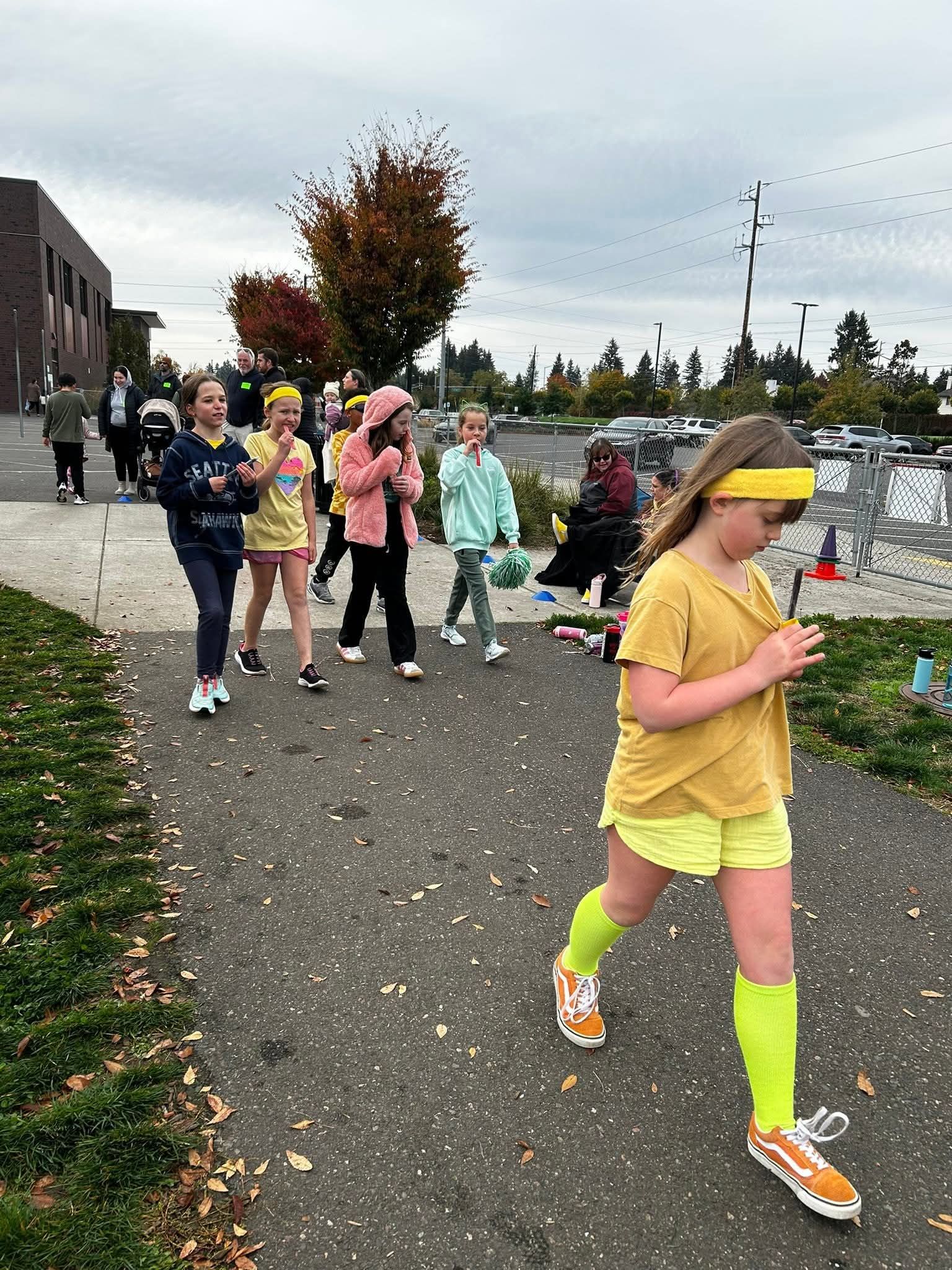 Group of kids walking on a path near a building, some wearing bright colors. Cloudy sky.