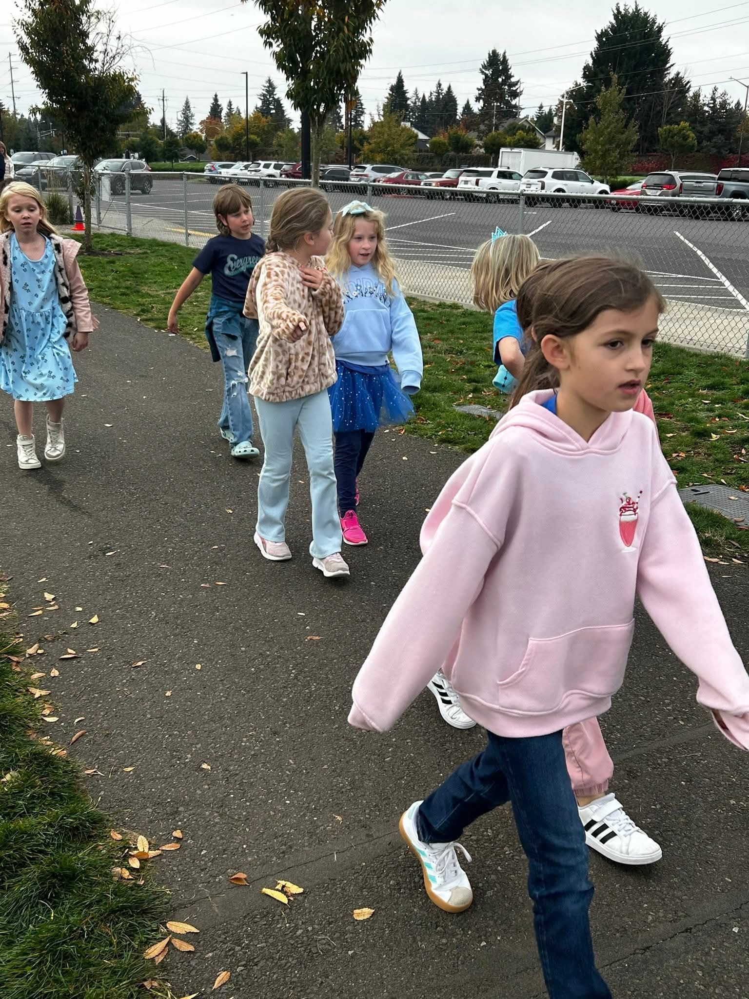 Children walking on a sidewalk near a parking lot, one wearing a pink hoodie.