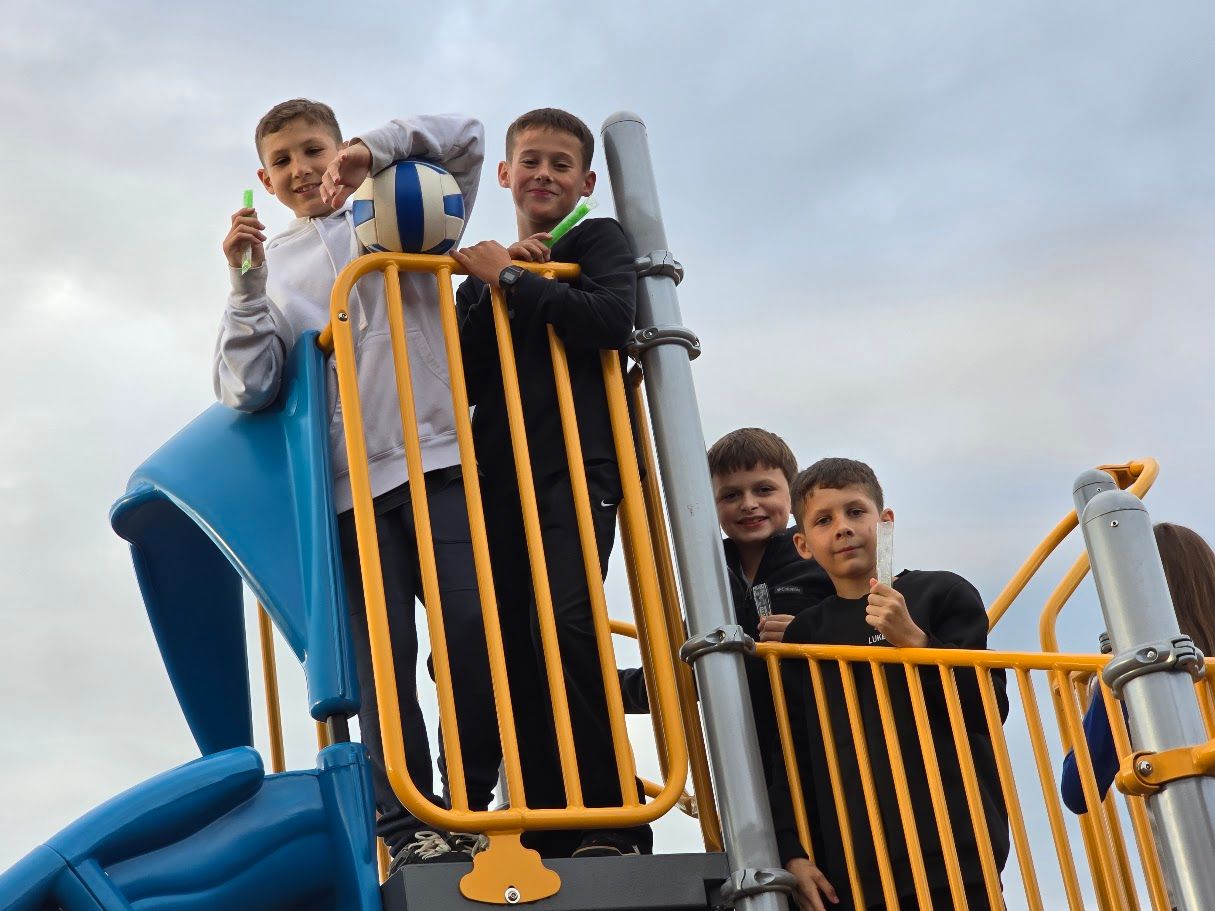 Four boys on a playground, posing and holding a soccer ball and a marker, with a cloudy sky.