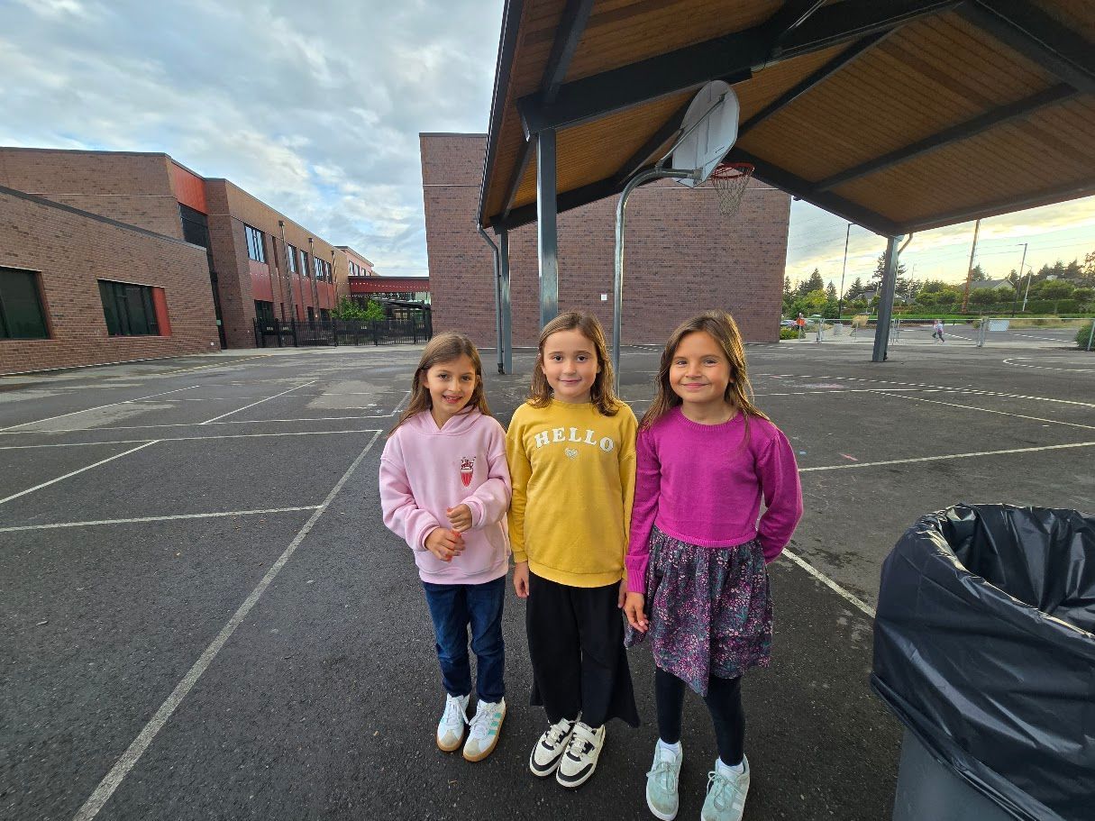 Three young girls standing in a schoolyard, smiling. Buildings and a canopy are in the background.