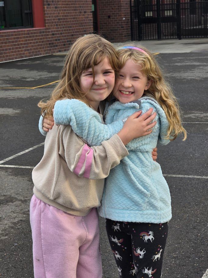 Two young girls hugging outdoors, smiling, wearing sweatshirts and leggings.
