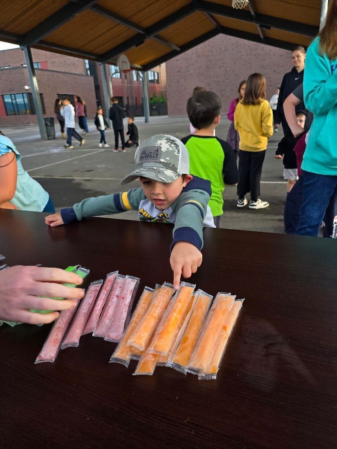 Boy pointing at orange ice pops on a table outside, other kids in the background.