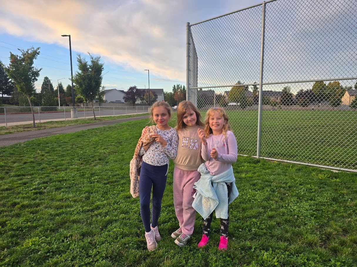 Three young girls smiling outdoors, standing on green grass near a chain link fence at dusk.