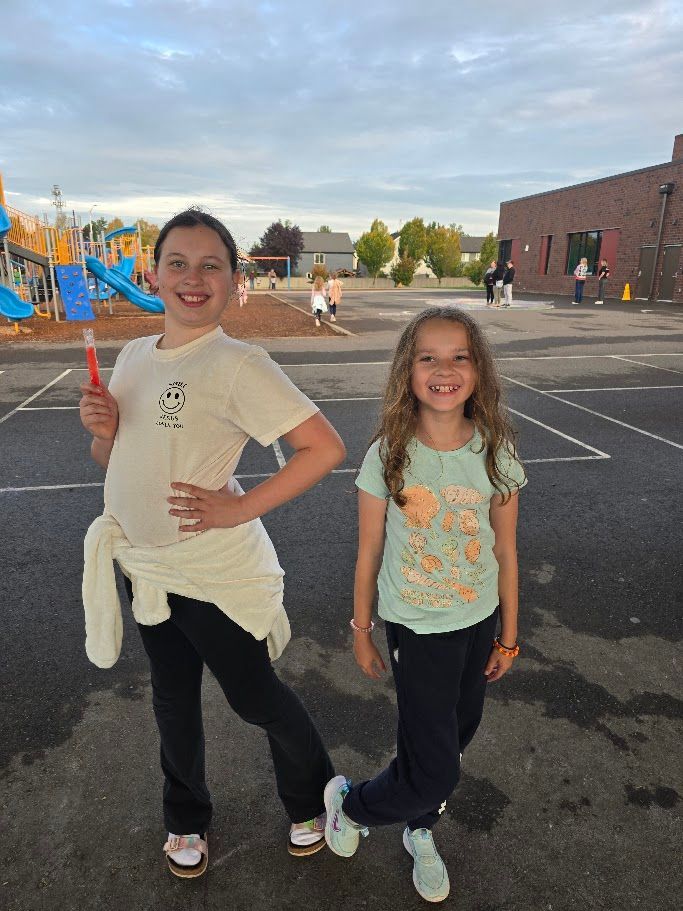 Two girls smiling, posing on a school playground. One holds an ice pop; the other has a foot raised.