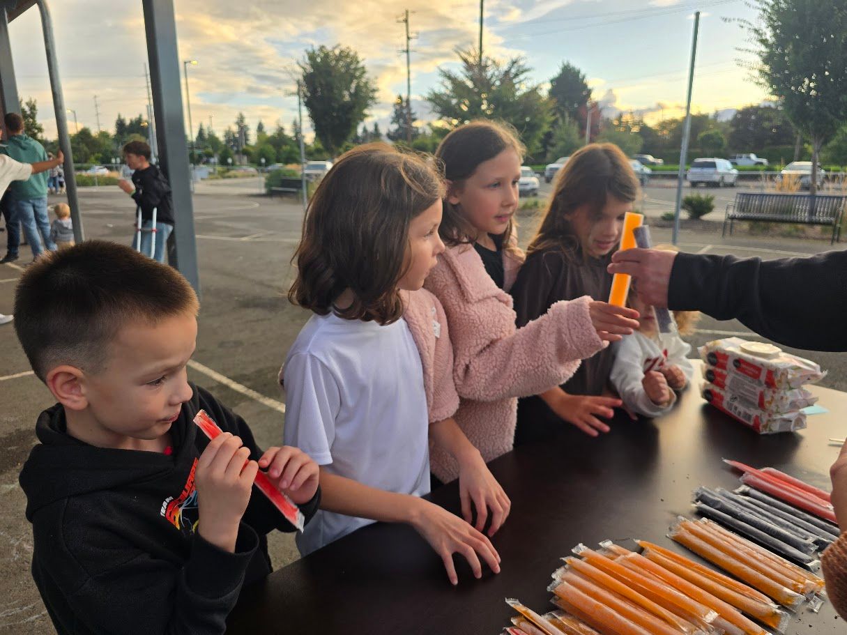 Children receiving frozen treats from a table outdoors.
