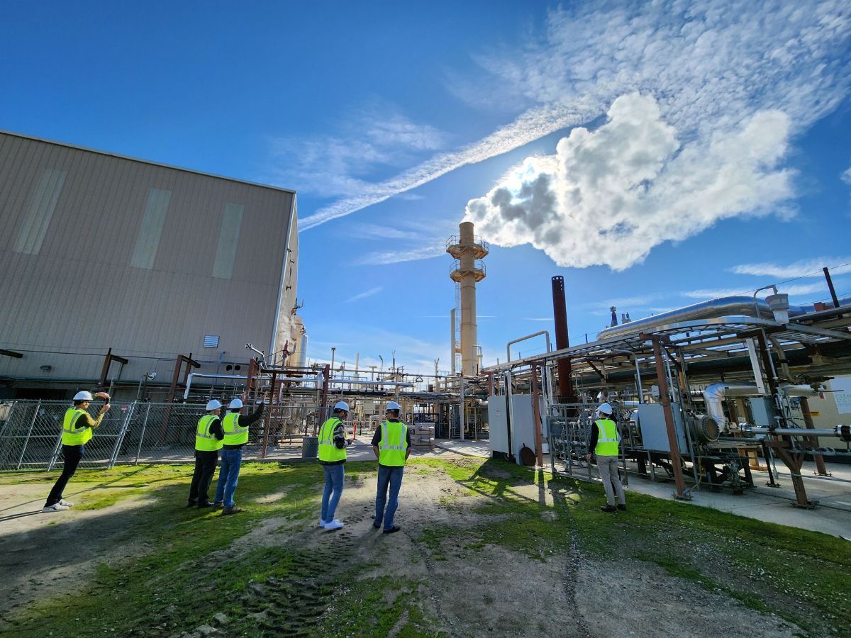 A group of people are standing in front of a factory with smoke coming out of it.