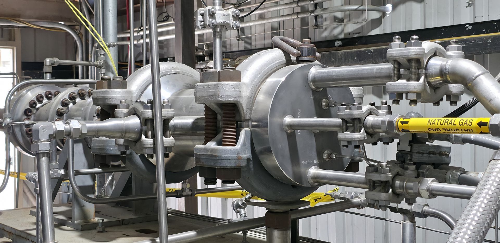 Industrial piping and turbine machinery in a factory, with silver pipes and yellow safety railings.