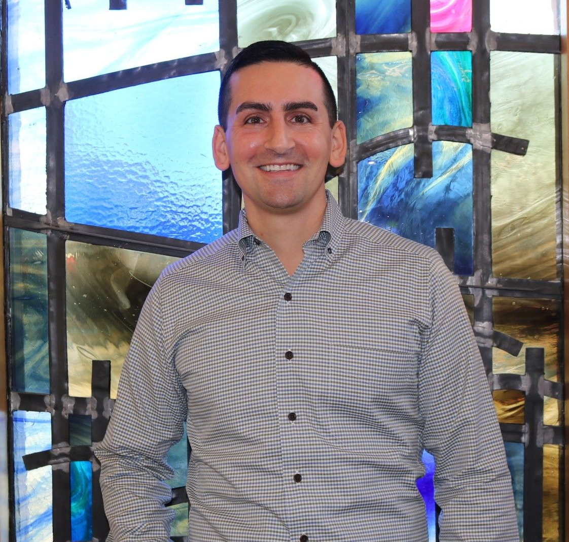 Man standing and smiling in a checkered shirt in front of a colorful stained-glass window