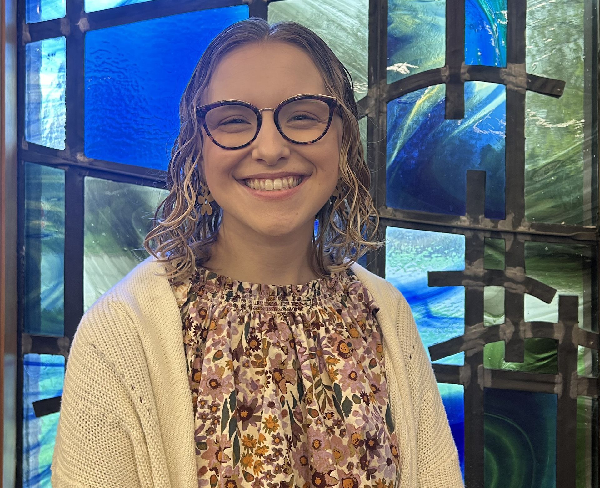 A woman wearing glasses is smiling in front of a stained glass window.
