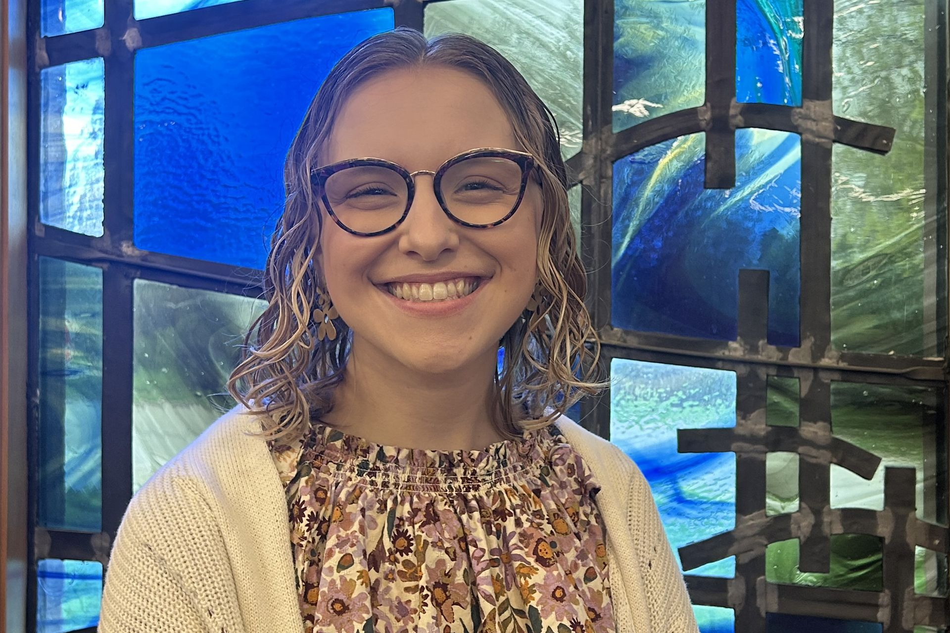 A woman wearing glasses is smiling in front of a stained glass window.