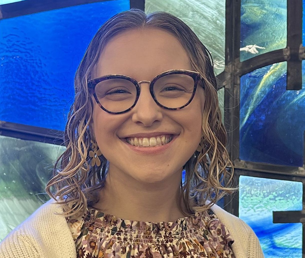 Smiling person with glasses in front of a blue stained-glass window