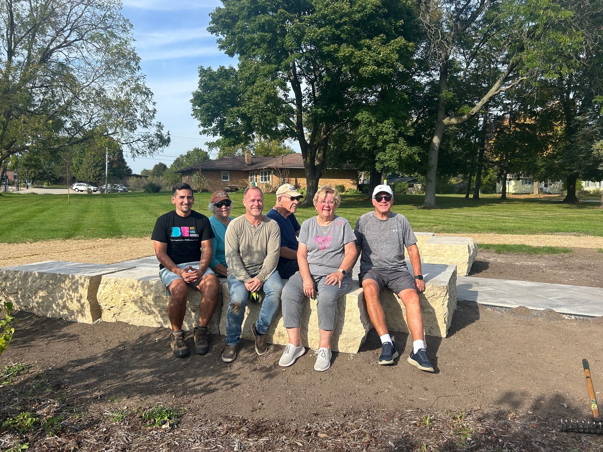Five people sit on a stone bench in a sunny park with trees and grass behind them.
