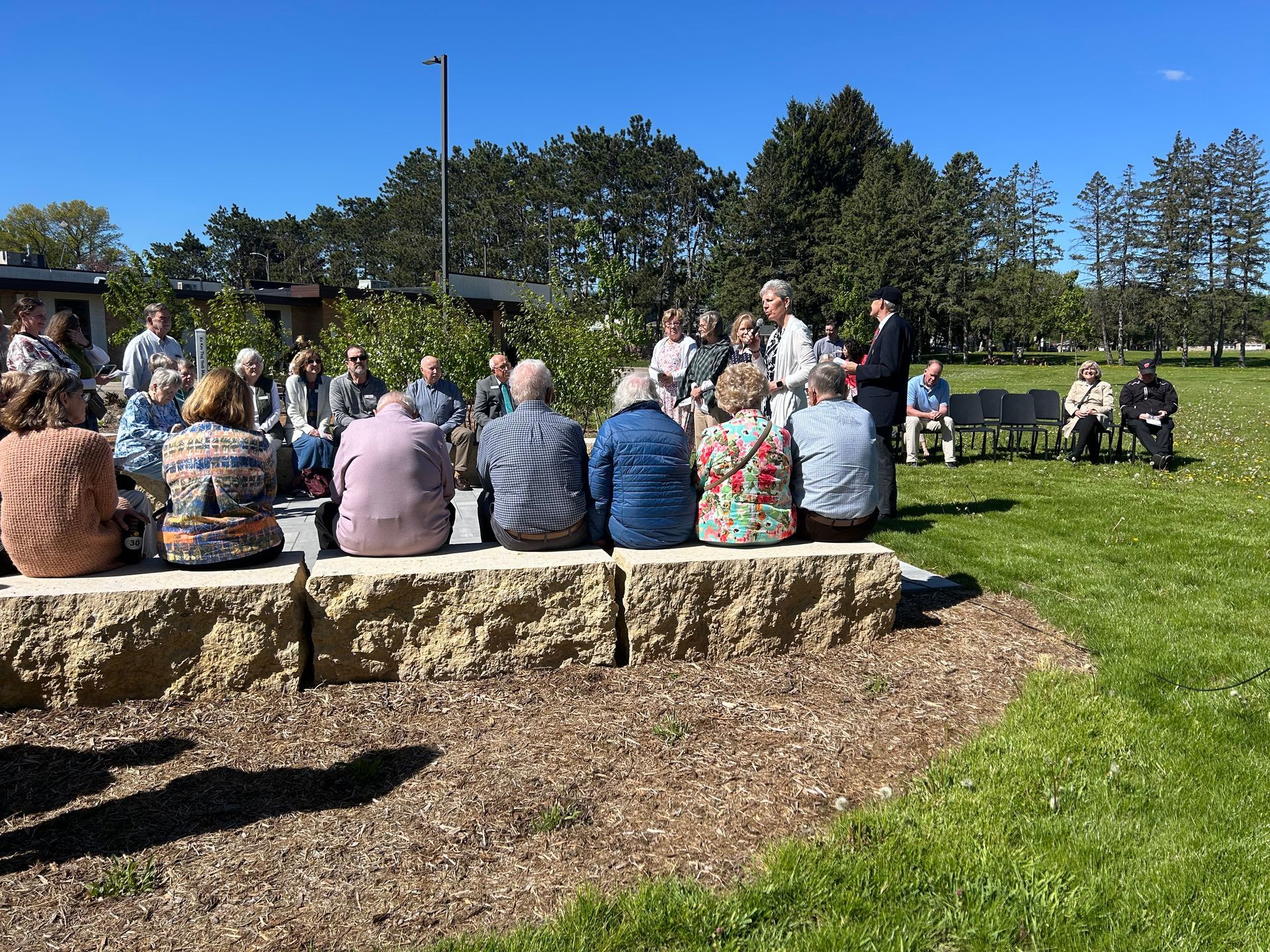 People seated on stone benches outdoors, listening to a gathering in a sunny park.