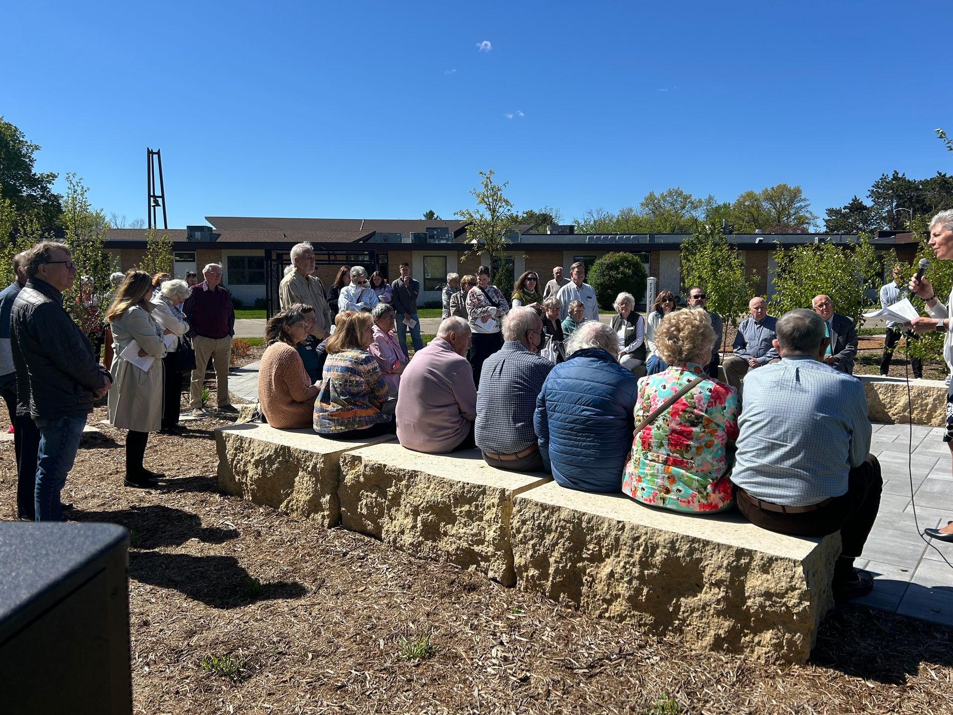 People seated on stone benches at an outdoor gathering in a sunny courtyard.