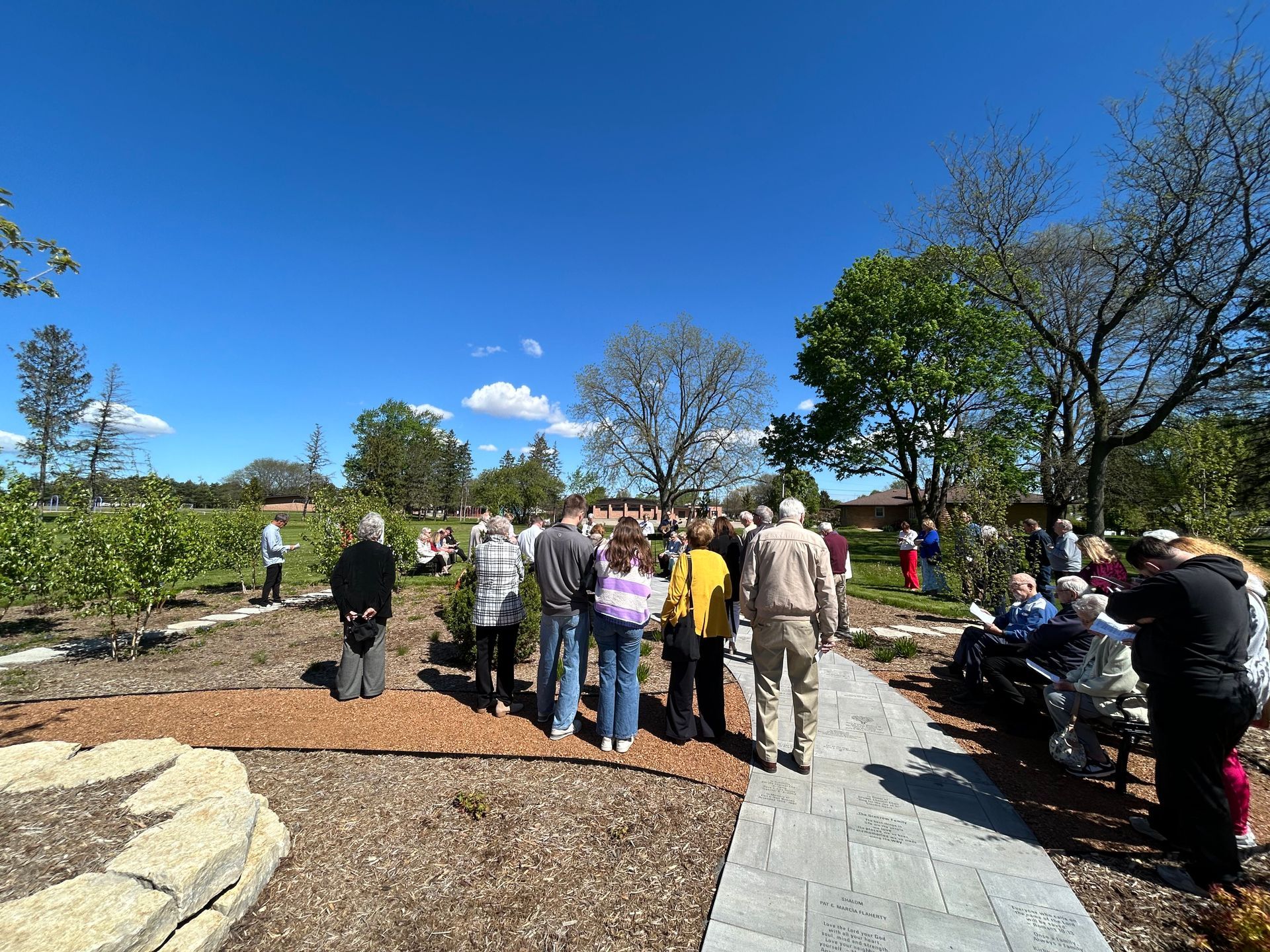 People gathered on a park path under a bright blue sky, with trees and a rocky landscape around them.
