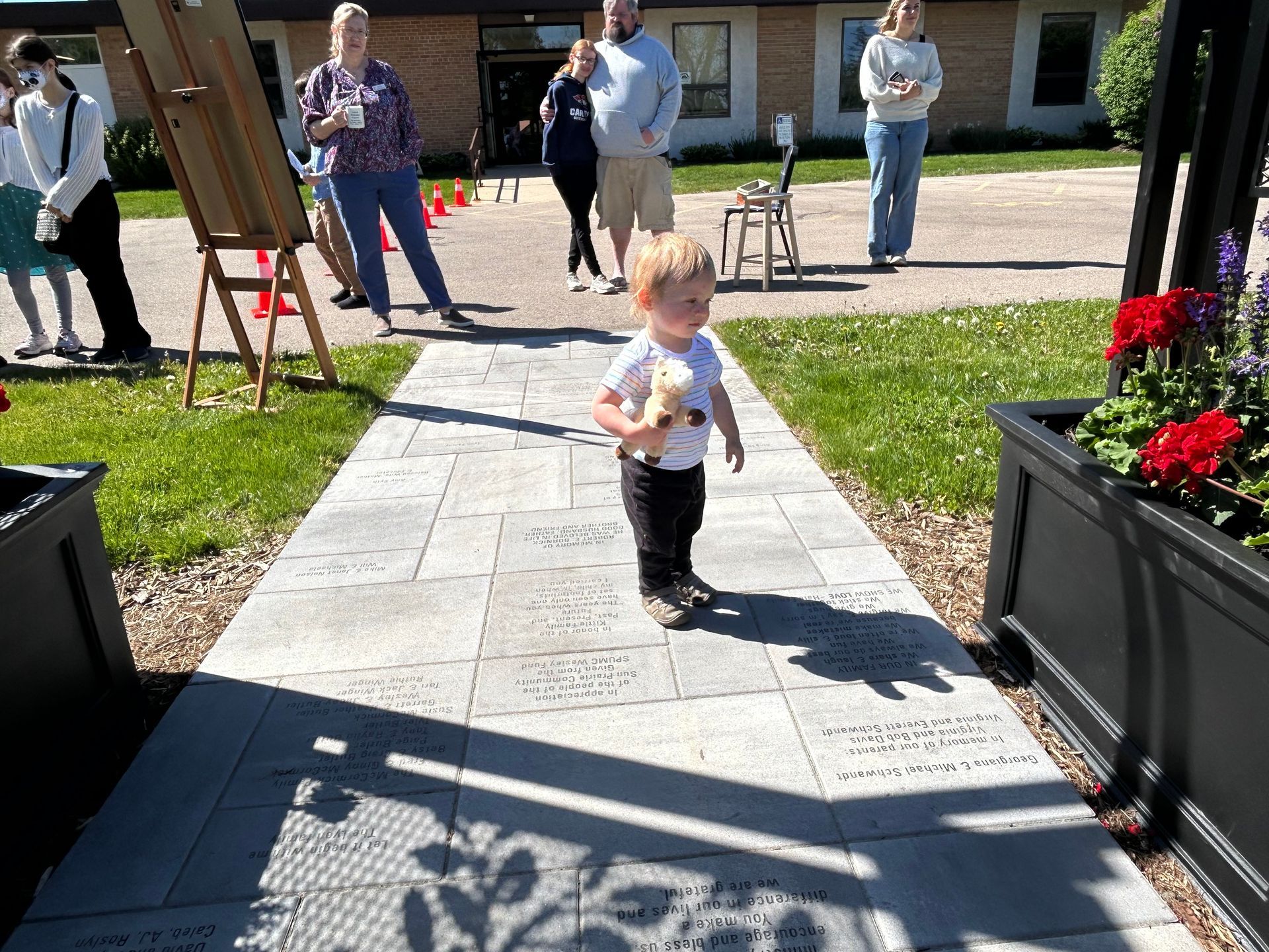 Toddler walking on a stone path toward a building, with adults and flowers in the background