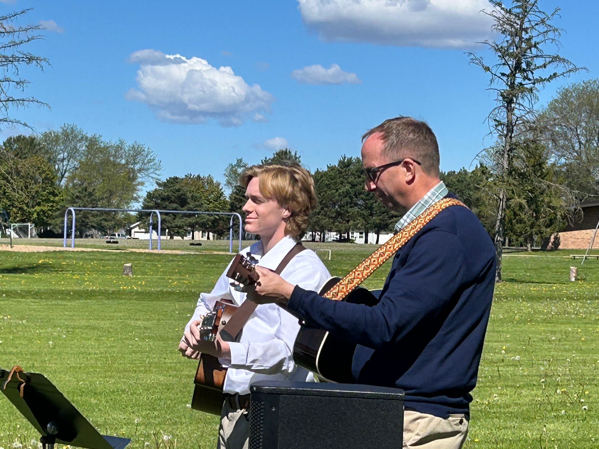 Two musicians playing guitar outdoors in a grassy park under a blue sky