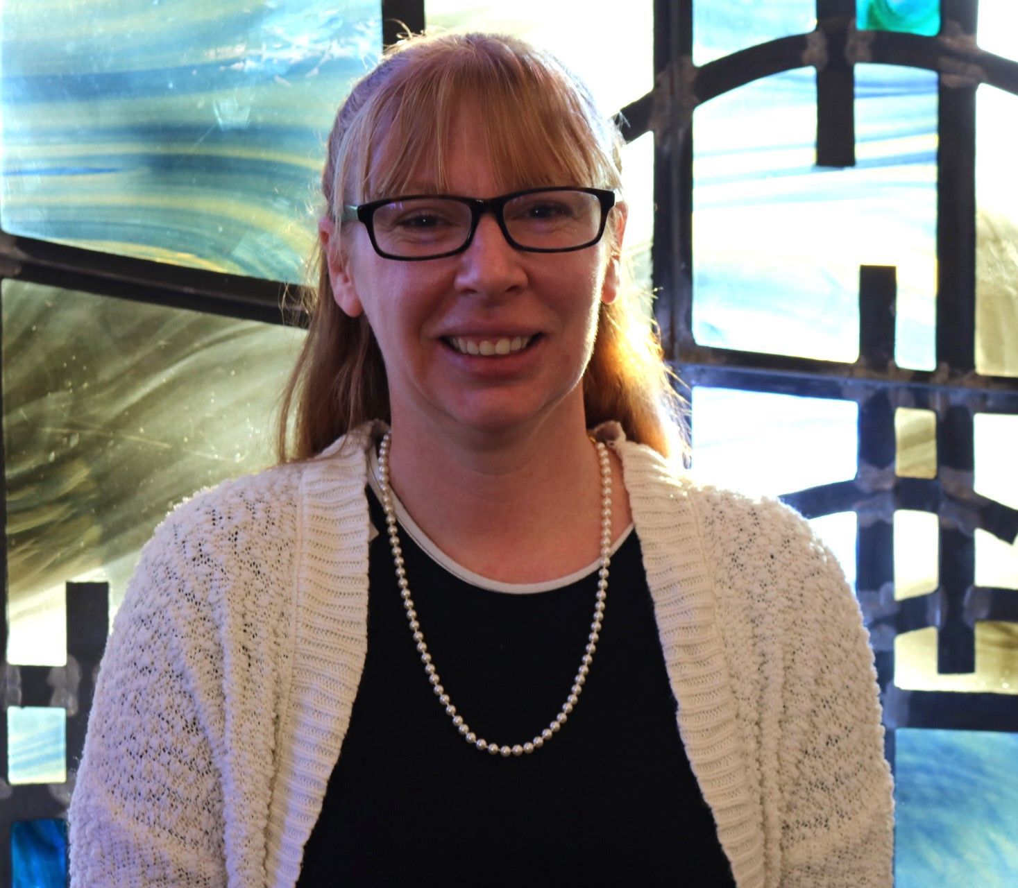 Smiling woman in glasses and white cardigan standing before a blue stained-glass window