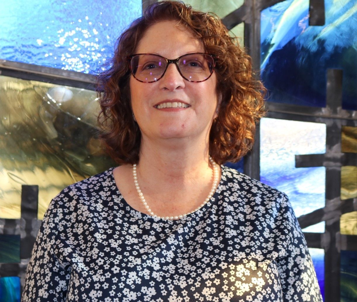 A woman wearing glasses and a pearl necklace is smiling in front of a stained glass window.