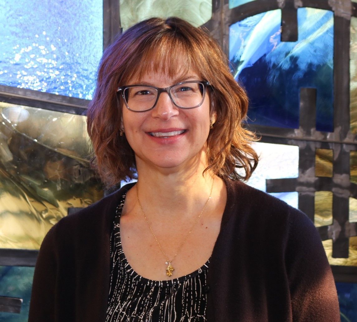 A woman wearing glasses is smiling in front of a stained glass window.