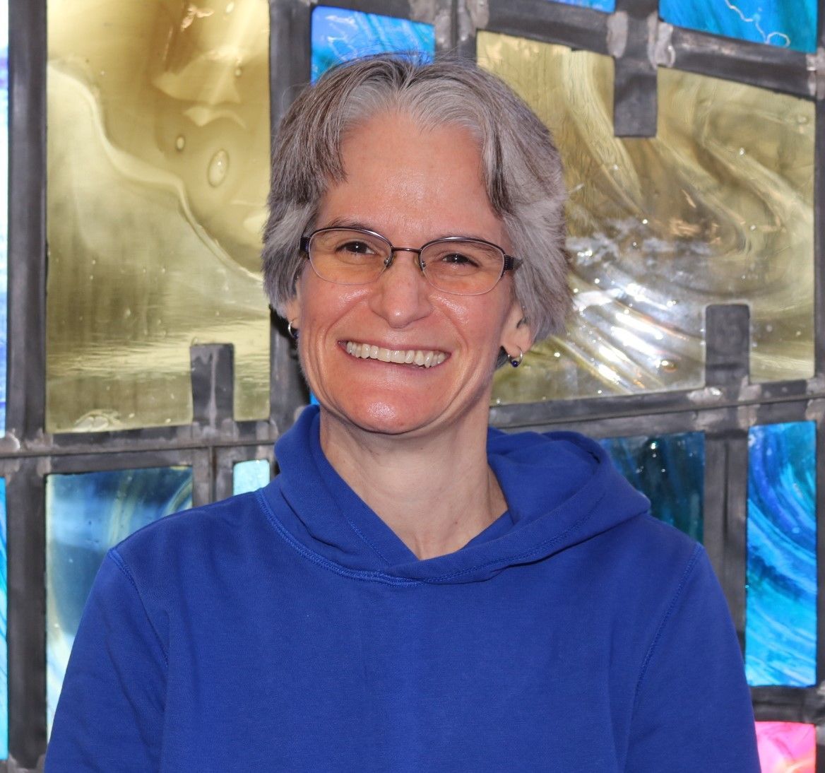 A woman wearing glasses and a blue sweater is smiling in front of a stained glass window.