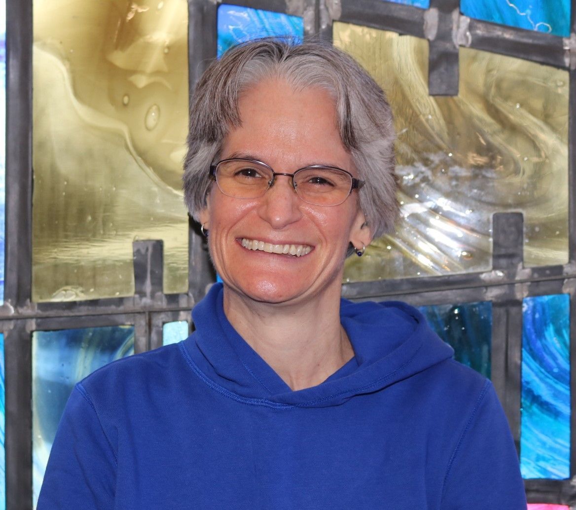 A woman wearing glasses and a blue sweater is smiling in front of a stained glass window