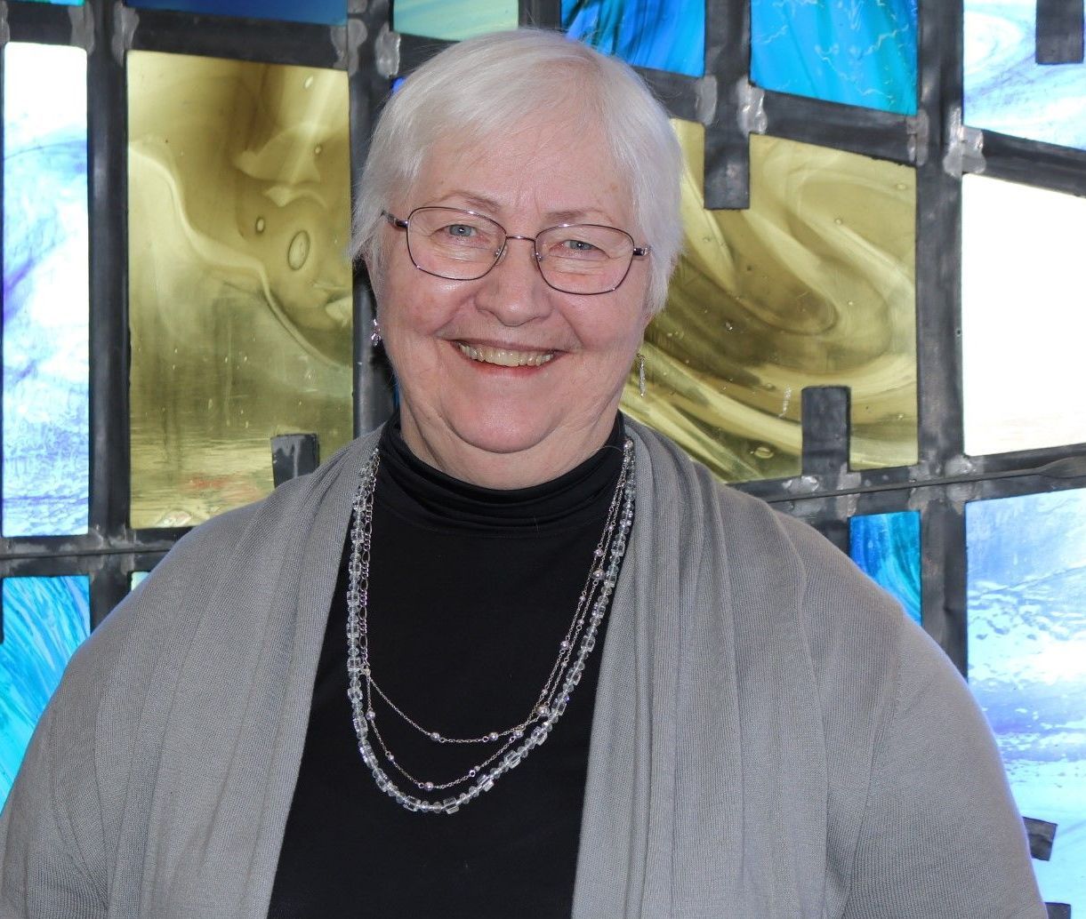 A woman wearing glasses and a necklace smiles in front of a stained glass window