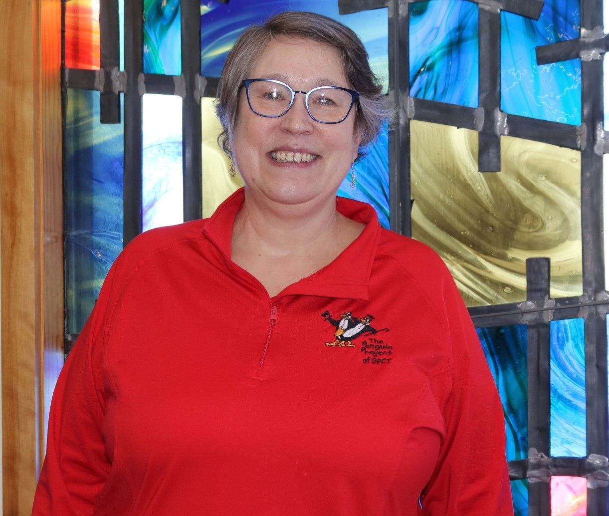 A woman wearing glasses and a red shirt is smiling in front of a stained glass window.