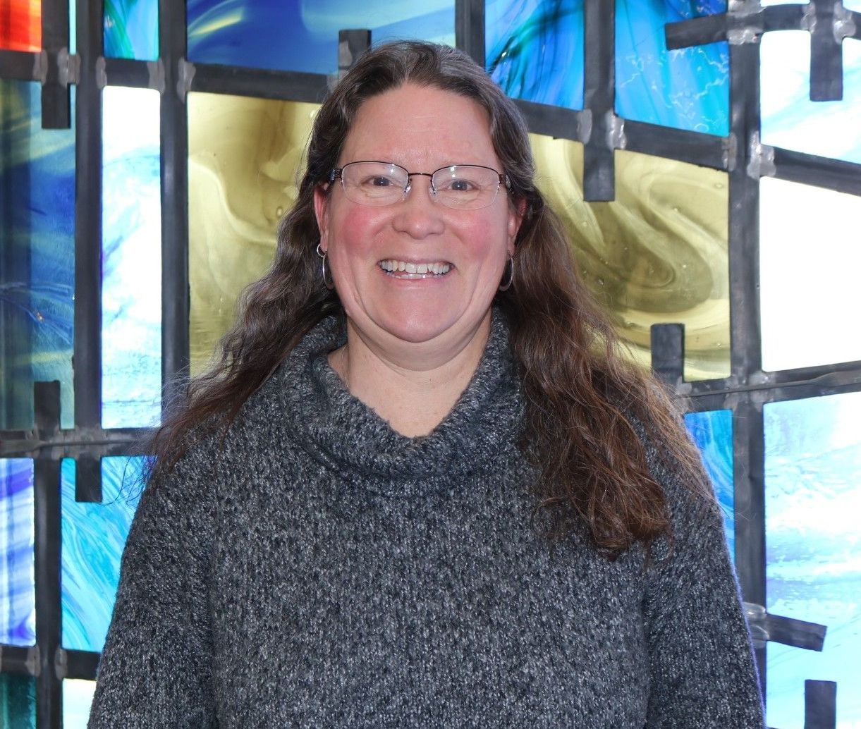 A woman wearing glasses and a grey sweater is smiling in front of a stained glass window.