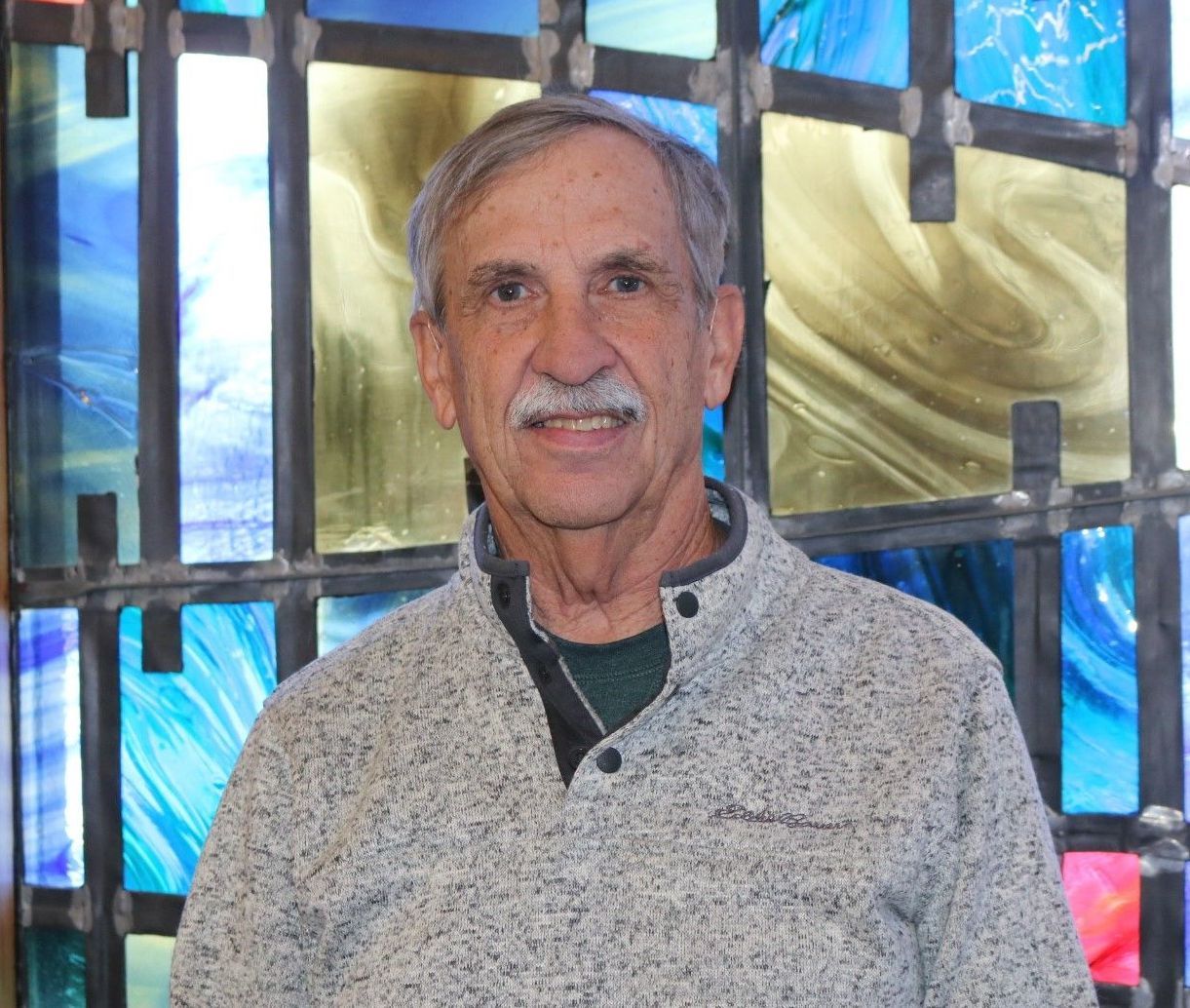 Smiling man in a gray sweater standing before a colorful stained-glass window