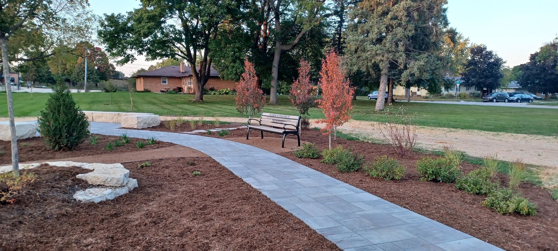 Curved concrete path through a landscaped park with mulch beds, trees, and benches under an overcast sky